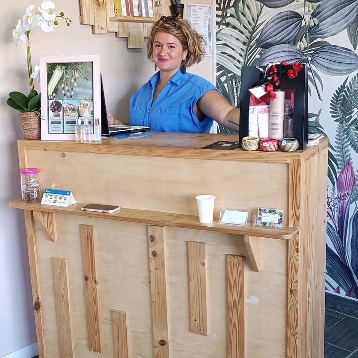 Émilie souriante derrière le comptoir en bois du salon, devant un papier peint à feuillages